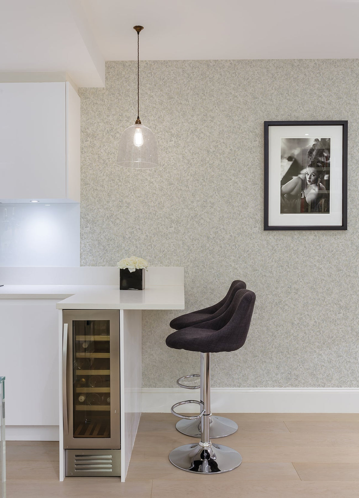Modern kitchen corner with two black bar stools by a white counter, pendant light above, a built-in wine fridge, a small flower arrangement, and a black-and-white framed photo on walls adorned with Hepworth Texture Wallpaper by York Wallcoverings.