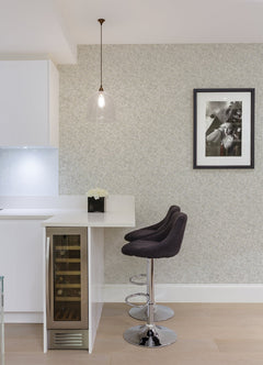 Modern kitchen corner with two black bar stools by a white counter, pendant light above, a built-in wine fridge, a small flower arrangement, and a black-and-white framed photo on walls adorned with Hepworth Texture Wallpaper by York Wallcoverings.