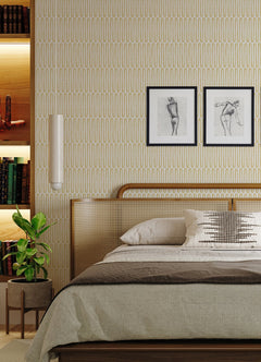 A cozy bedroom featuring a cane headboard, two framed sketches, a potted plant, and shelves with books and decor. The bed is neatly made with neutral bedding and a decorative pillow, while the walls are adorned in York Wallcoverings’ Lars Retro Wave Wallpaper.