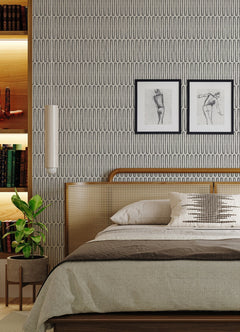 A modern bedroom featuring York Wallcoverings’ Lars Retro Wave Wallpaper, two framed sketches displayed above a cane headboard, neutral bedding accented with a decorative pillow, a potted plant, and built-in shelves styled with books and art objects.