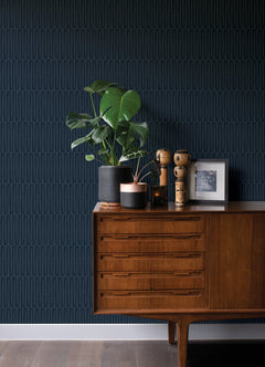 A wooden sideboard with potted plants, small sculptures, and a framed photo is set against a dark blue patterned wall featuring Lars Retro Wave Wallpaper by York Wallcoverings, which displays vertical oval shapes. The floor is medium-brown wood.