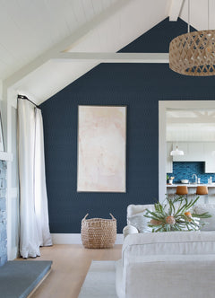 A modern living room featuring a dark blue Lars Retro Wave Wallpaper by York Wallcoverings as an accent wall, complemented by light abstract art, a beige sofa, a woven basket, a large plant, and a view into a bright kitchen with bar stools and a blue backsplash.