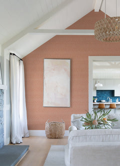 A modern living room featuring a Lars Retro Wave Wallpaper by York Wallcoverings as a peach-patterned accent wall, complemented by abstract art, light furnishings, a woven basket, and an open kitchen with a blue backsplash and wooden stools.