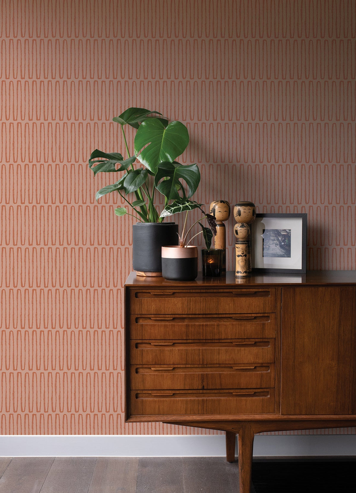 A mid-century modern wooden sideboard adorned with potted plants, wooden figurines, and a framed photo sits against a wall covered in York Wallcoverings’ Lars Retro Wave Wallpaper featuring vertical oval patterns.