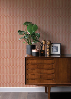 A mid-century modern wooden sideboard adorned with potted plants, wooden figurines, and a framed photo sits against a wall covered in York Wallcoverings’ Lars Retro Wave Wallpaper featuring vertical oval patterns.