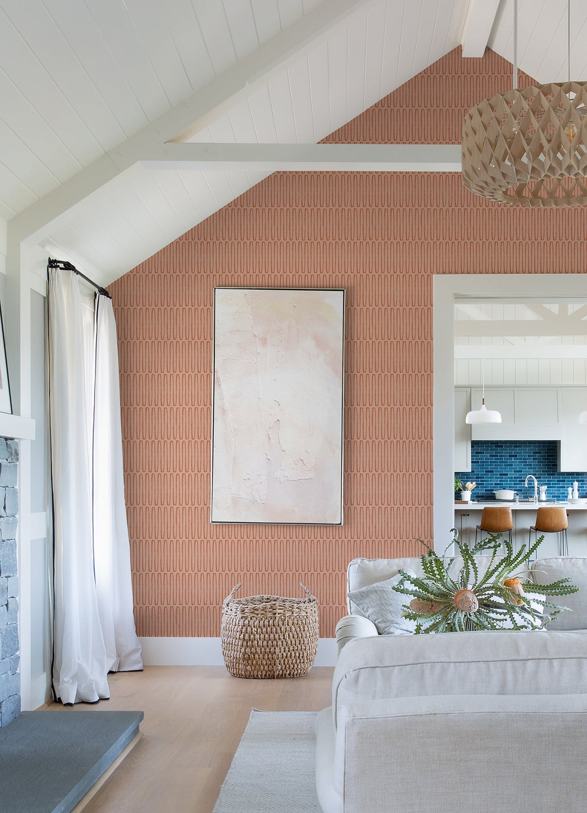 A cozy living room featuring a Lars Retro Wave Wallpaper by York Wallcoverings as a patterned accent wall, abstract art, light furniture, a woven basket, and a view into a modern kitchen with blue backsplash and wooden stools.