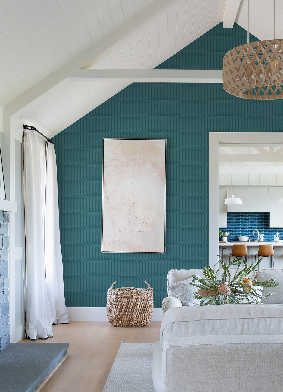Modern living room with a vaulted white ceiling, an accent wall featuring York Wallcoverings’ Lars Retro Wave Wallpaper, abstract art, woven basket, light sofa, potted plant, and an open view into a kitchen with a blue tile backsplash and wooden stools at the counter.