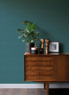 Against a dark floor, a wooden sideboard is styled with potted plants, wooden figurines, and framed art, set in front of a teal patterned wall adorned with York Wallcoverings’ Lars Retro Wave Wallpaper.