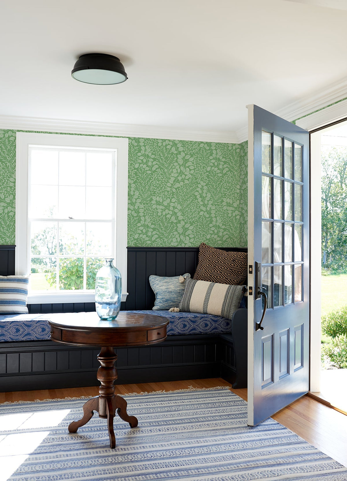 A cozy room featuring York Wallcoverings’ Elin Berry Botanical Wallpaper in green, a built-in bench with striped and patterned pillows, a round wooden table topped with a glass vase, a striped rug, and an open door letting in sunlight.