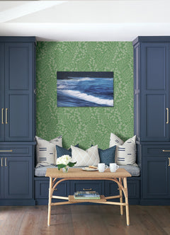 A cozy bench nook with blue cabinets, Elin Berry Botanical Wallpaper by York Wallcoverings in a green floral pattern, decorative pillows, a wicker table topped with flowers, tea cups, and ocean wave wall art. A wooden floor completes the inviting space.