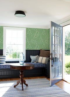 A cozy room with a blue door open to the outside features a round wooden table topped with a glass vase, built-in bench seating with cushions, York Wallcoverings’ Elin Berry Botanical Wallpaper in green on the walls, and a striped rug on the floor.