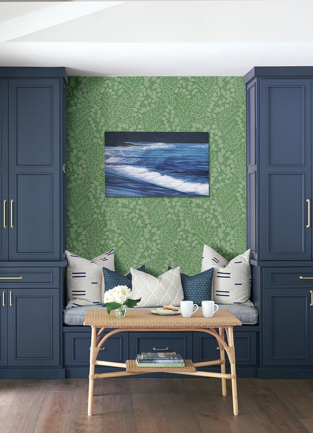 Blue built-in cabinets frame a cozy bench with patterned pillows, a wicker table with mugs, a vase of flowers, and Elin Berry Botanical Wallpaper by York Wallcoverings in lush green leafy patterns. An ocean wave painting hangs above the bench.