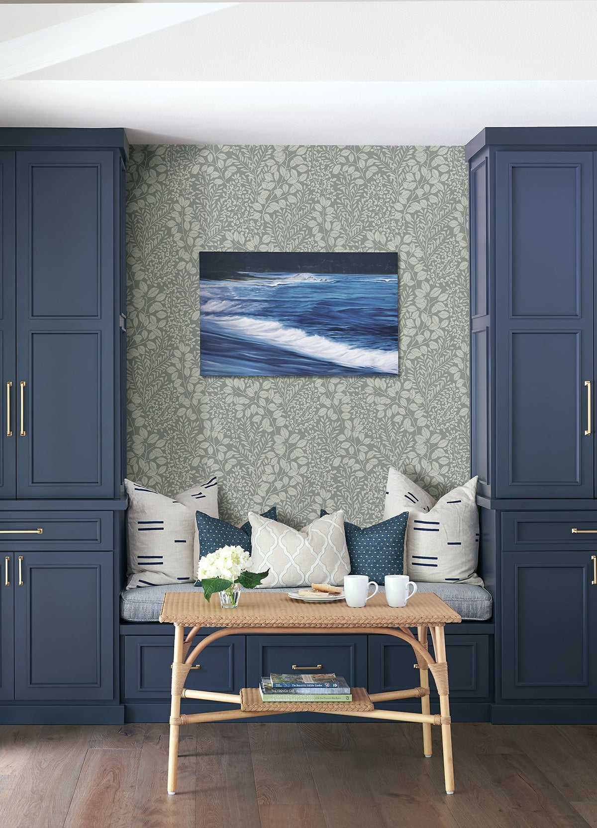 Cozy nook with blue cabinets, a cushioned bench adorned with patterned pillows, a wicker table set with tea and flowers, and a seascape painting on walls decorated in Elin Berry Botanical Wallpaper by York Wallcoverings.