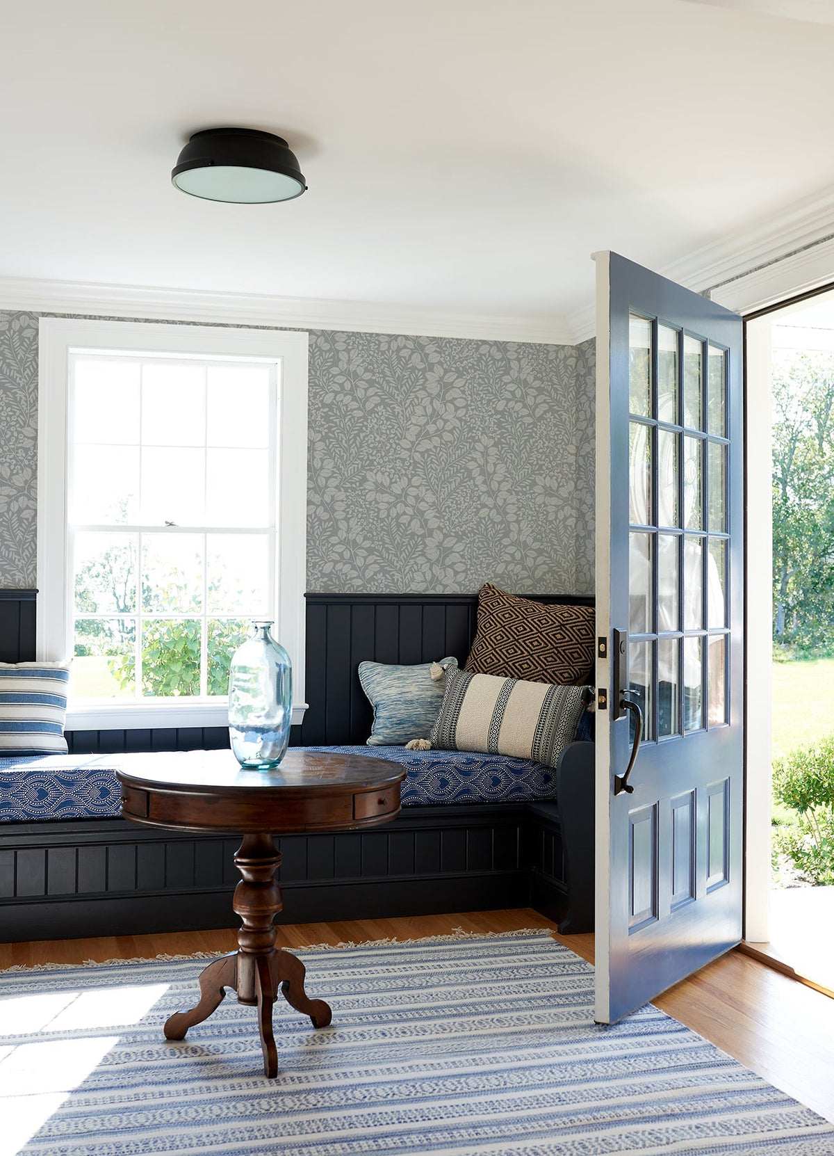 A sunlit room features a cozy corner bench adorned with patterned blue cushions, a round wooden table topped with a glass vase, and walls decorated in Elin Berry Botanical Wallpaper by York Wallcoverings. An open blue door leads outside, and a striped rug lies over the wooden floor.