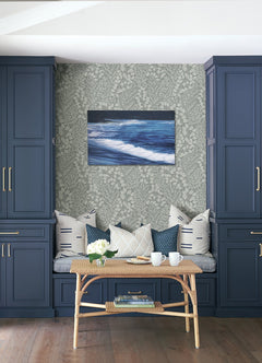 A cozy bench with patterned pillows sits between blue cabinets. In front, a rattan table holds white mugs, a teapot, and flowers. On the wall, ocean artwork is displayed against Elin Berry Botanical Wallpaper by York Wallcoverings.