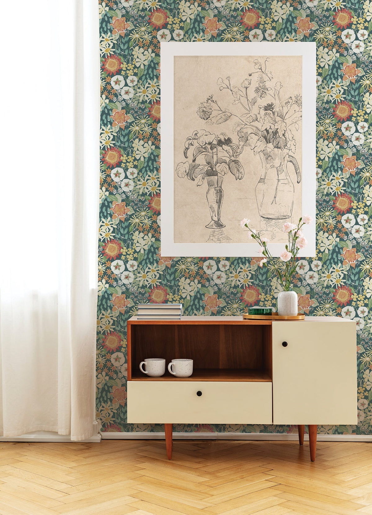 A sideboard with two mugs sits against a wall adorned in Karina Wildflower Garden Wallpaper by York Wallcoverings. Above is a framed black-and-white flower sketch, while a vase with pink flowers rests on the sideboard; white curtains hang to the left.
