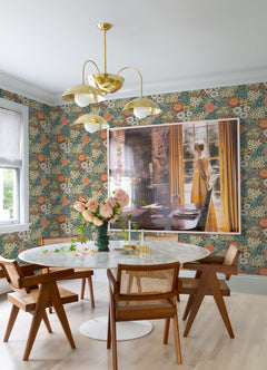 Dining area featuring a round white table, wooden chairs, a gold chandelier, vase of flowers, and large framed photo of a woman in a yellow dress by sunlit curtains—accented with York Wallcoverings’ Karina Wildflower Garden Wallpaper.