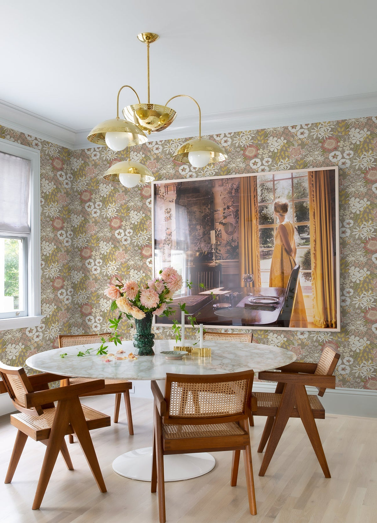 A dining room featuring York Wallcoverings’ Karina Wildflower Garden Wallpaper, a round white marble table, wooden chairs, a vase of flowers, gold chandelier, and a large framed photo of a person set against yellow curtains on the wall.