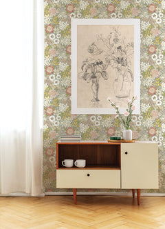 A mid-century cabinet with two white mugs and a vase holding a pink flower sits against a wall adorned with York Wallcoverings’ Karina Wildflower Garden Wallpaper, topped by a large sketch of flowers in vases above. Light wood flooring and sheer curtains complete the cozy scene.