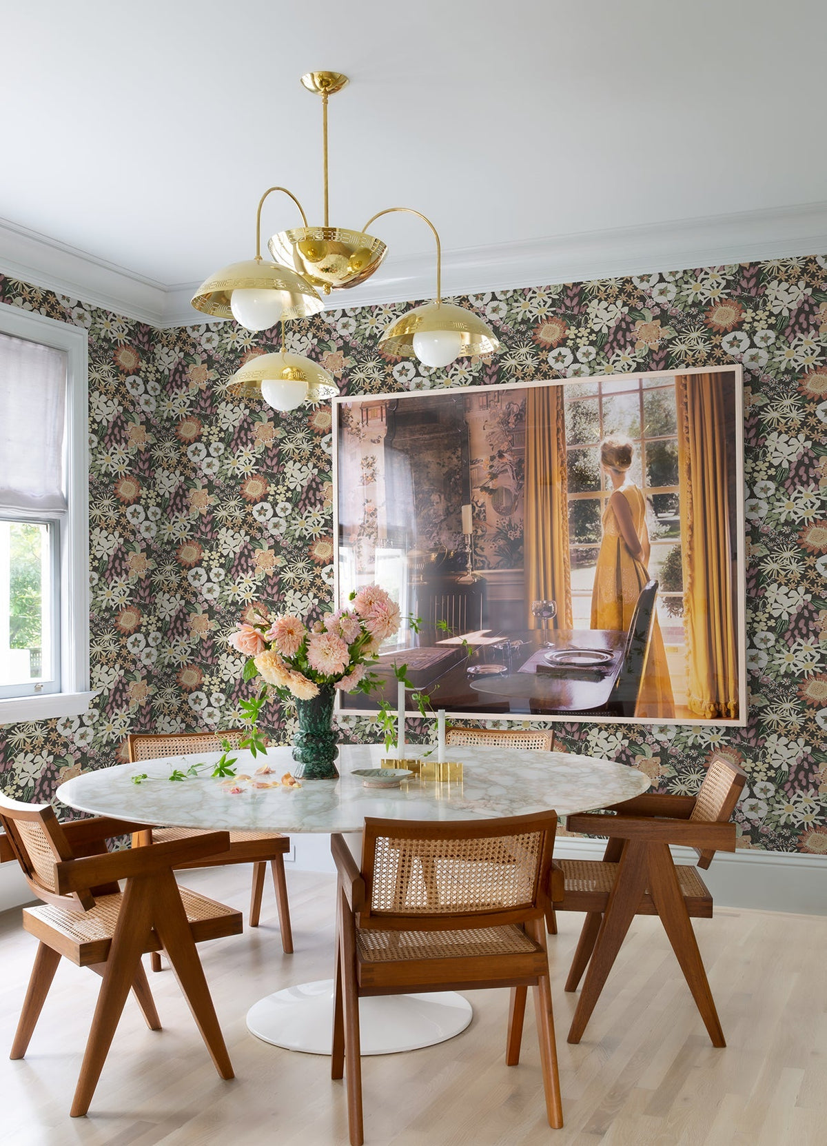A dining area with a round white table, wooden chairs, a vase of pink flowers, a gold chandelier, wall art of a woman in a yellow dress looking out a window, and the Karina Wildflower Garden Wallpaper by York Wallcoverings.