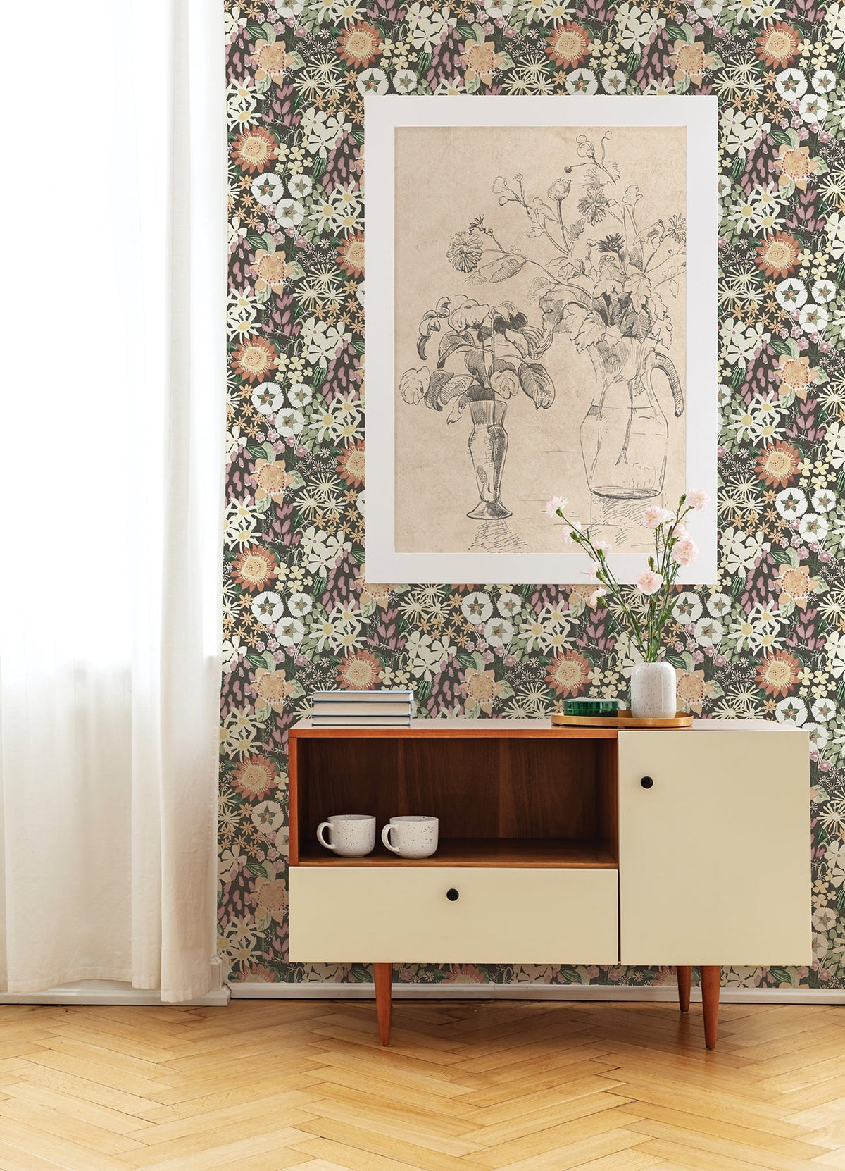 A room adorned with Karina Wildflower Garden Wallpaper by York Wallcoverings features a beige sideboard, two white mugs, a glass vase filled with flowers, a large floral sketch on the wall, and sheer white curtains.