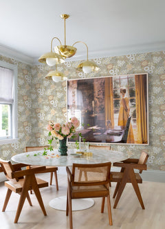 A bright dining room featuring a round white table, four wooden chairs, a vase of flowers, a gold chandelier, and large art of a woman in a yellow dress by a window with yellow curtains, with the walls adorned in Karina Wildflower Garden Wallpaper by York Wallcoverings.