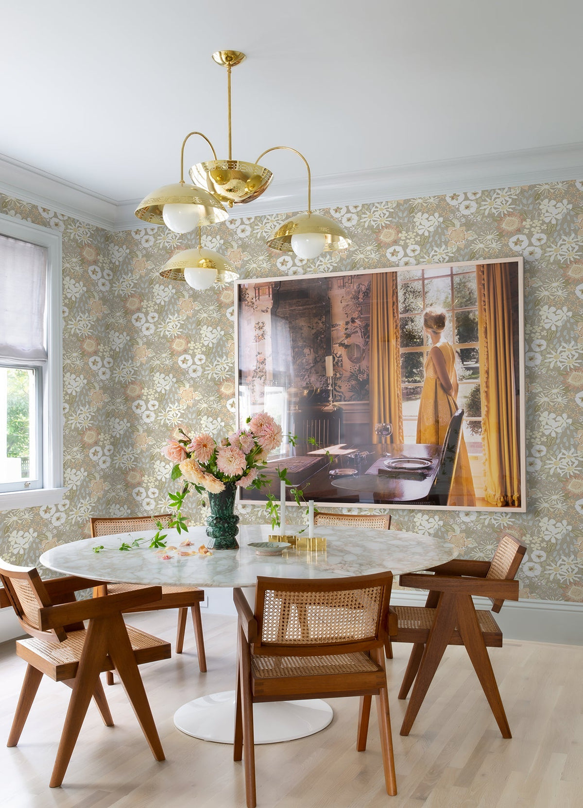 A dining room featuring a round marble table and wooden chairs, adorned with York Wallcoverings’ Karina Wildflower Garden Wallpaper, a gold chandelier, and a large framed photo of a woman in a yellow dress by a window. Pink flowers and glassware are arranged on the table.