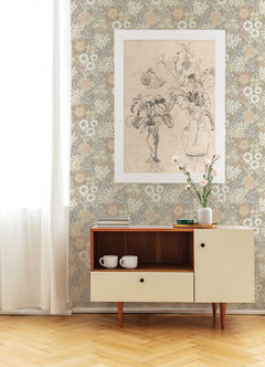 A mid-century modern cabinet holds two white mugs and a vase with pink flowers. Behind it, Karina Wildflower Garden Wallpaper by York Wallcoverings and a large sketch of flowers in vases decorate the wall. Sunlight streams through a sheer curtain.