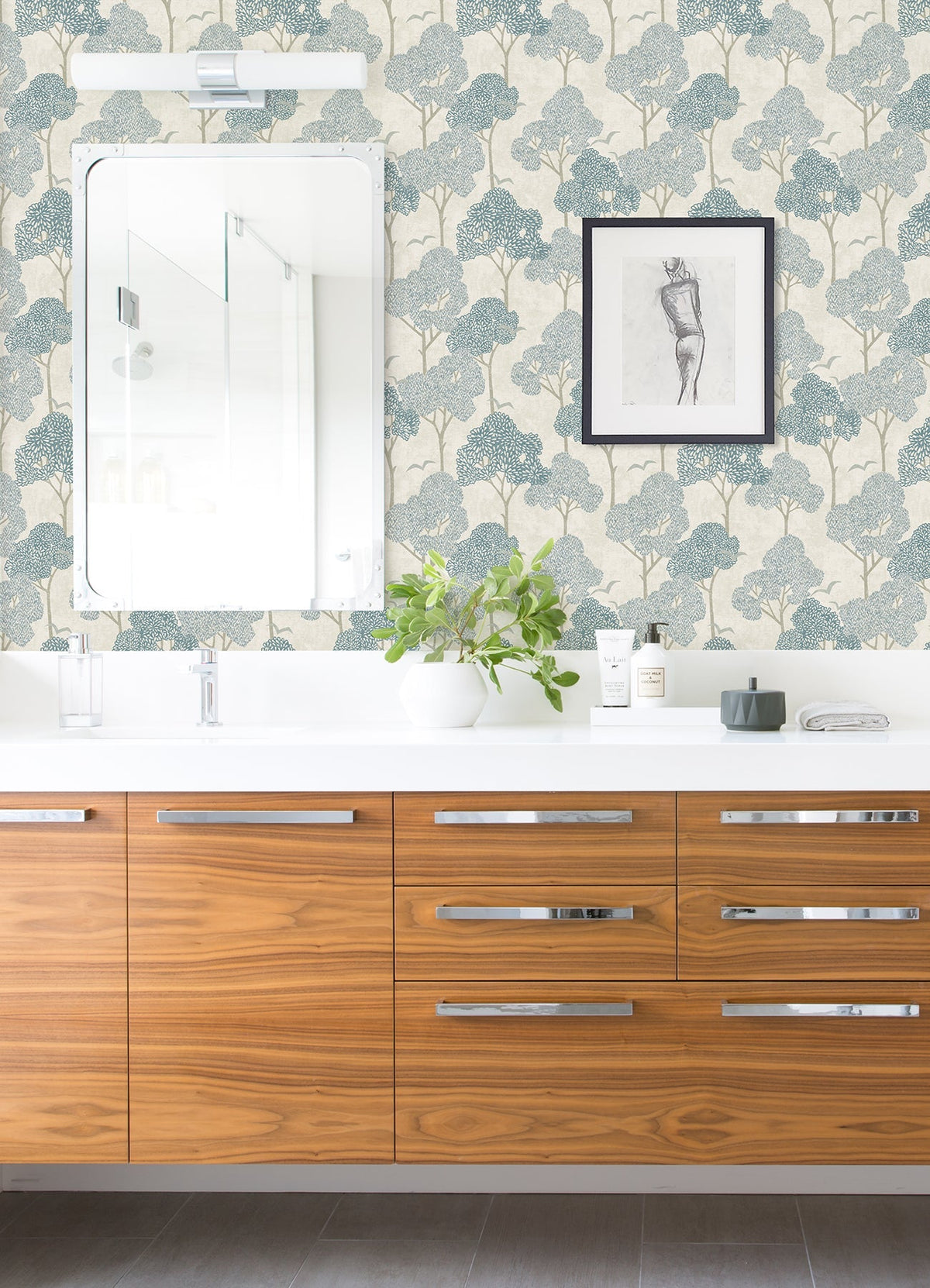 Bathroom with wooden cabinets, a white countertop, a mirror, and a plant in a glass vase. The wall features York Wallcoverings’ Lykke Textured Tree Wallpaper in blue leafy tones and is accented by a framed sketch of a figure. Toiletries sit beside the sink.