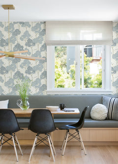 A dining nook with black chairs, a wooden table, a built-in bench with gray cushions, and a window features York Wallcoverings’ Lykke Textured Tree Wallpaper on the wall. A vase of flowers sits on the table.