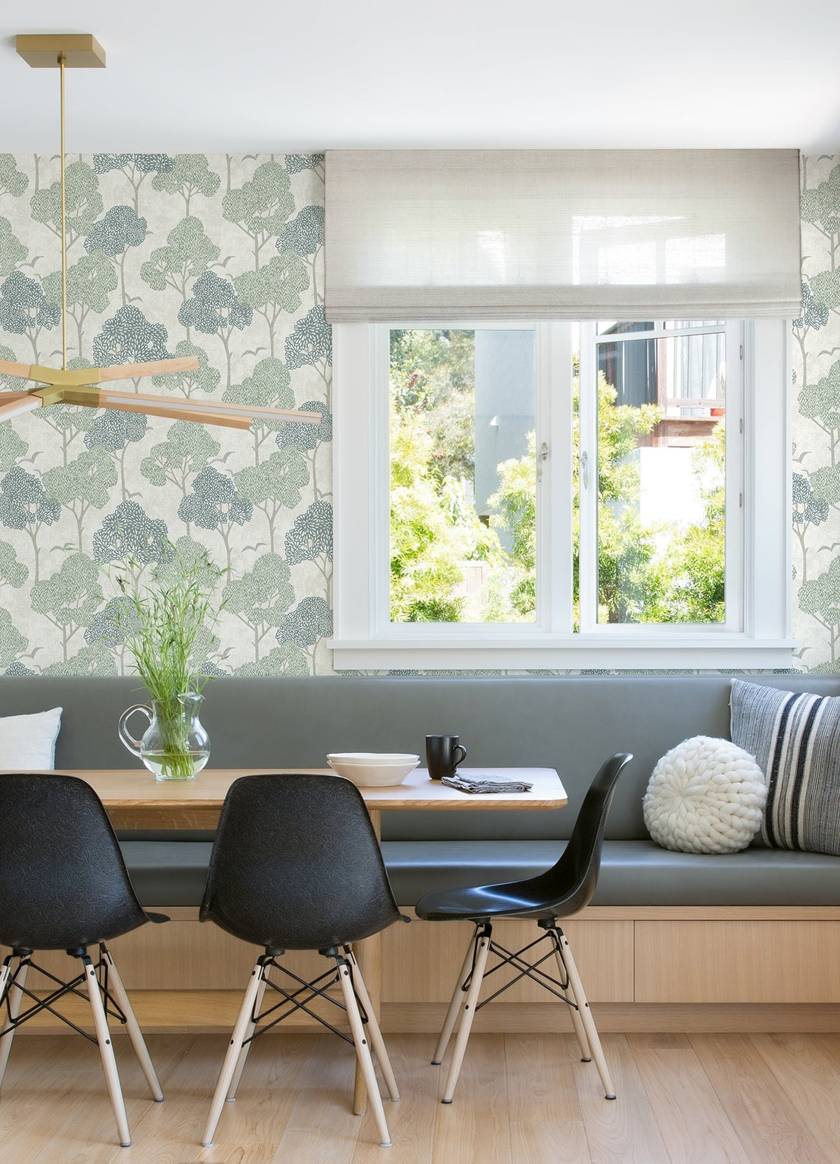 A modern dining nook featuring black chairs, a wooden table, light bench seating, a window overlooking greenery, minimalist decor, and walls adorned with York Wallcoverings’ Lykke Textured Tree Wallpaper.