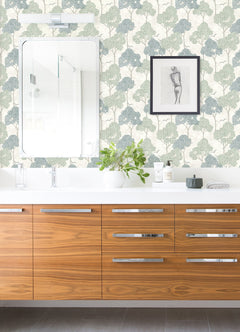 Modern bathroom with a wooden vanity, white countertop, mirror, plant, toiletries, and framed art on a feature wall adorned with York Wallcoverings’ Lykke Textured Tree Wallpaper in blue and green patterns.