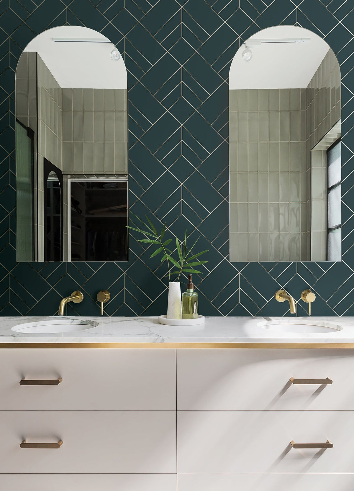 Double bathroom vanity with brass fixtures, white countertop, two modern mirrors, York Wallcoverings Sander Geometric Wallpaper on the wall, and a white vase with green leaves between the sinks. Light reflects from a window on the right.