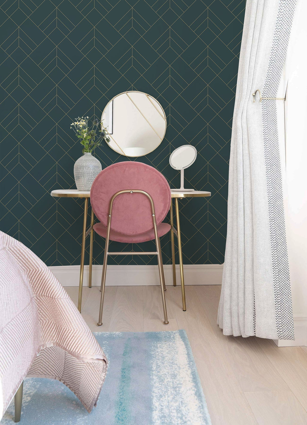 A vanity with a round mirror, white vase with flowers, and a pink chair stands against a wall covered in Sander Geometric Wallpaper by York Wallcoverings. Light curtains and a pastel rug are also visible.