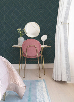 A small vanity with a pink chair, gold legs, round mirrors, and a vase of flowers sits against the Sander Geometric Wallpaper by York Wallcoverings. Cream curtains and a bed with a striped blanket are also visible.