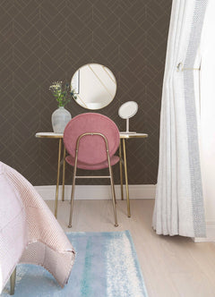 A small vanity table with a round mirror, pink chair, vase of flowers, and table mirror stands against a wall covered in York Wallcoverings’ Sander Geometric Wallpaper. Light curtains and part of a bed and rug are visible in the foreground.