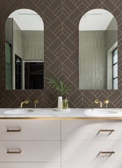 Modern bathroom with double sinks, brass fixtures, white countertop, and taupe drawers. Two arched mirrors reflect light green walls adorned with Sander Geometric Wallpaper by York Wallcoverings. A vase with green leaves sits between the sinks. Geometric brown tile backsplash.