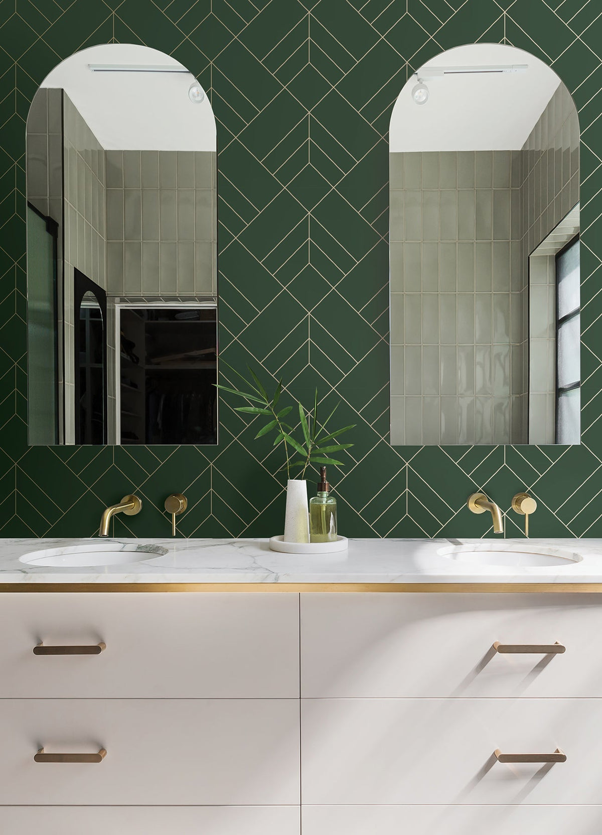 Modern bathroom featuring two arched mirrors, a green Sander Geometric Wallpaper by York Wallcoverings on the wall, double sinks with brass faucets, and a white vanity adorned with gold accents. A vase with green leaves is placed between the sinks.