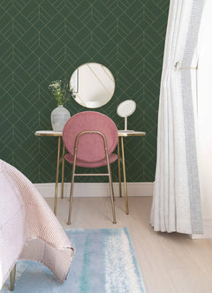 A bright, modern room features a vanity with a pink chair, round mirror, small makeup mirror, and a vase of flowers, set against York Wallcoverings’ Sander Geometric Wallpaper in green. A bed and curtain are partly visible in the background.