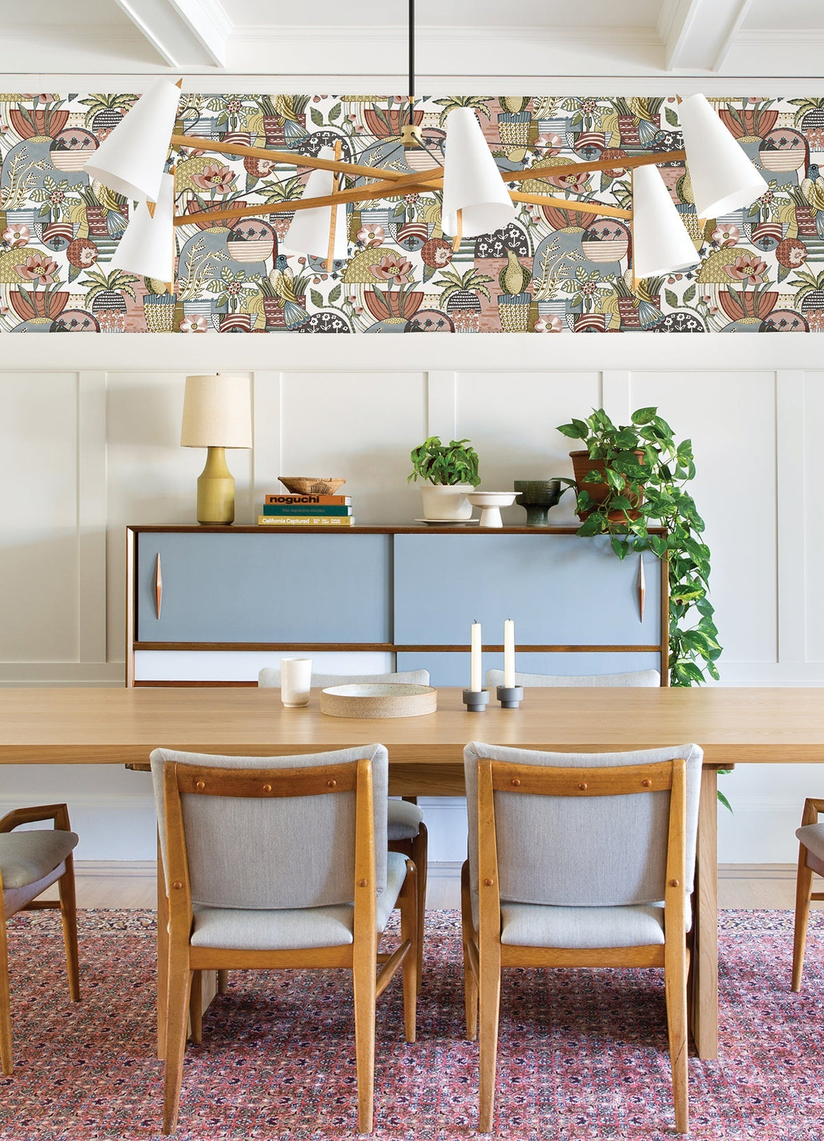 A modern dining room featuring a wooden table, four gray chairs, a mid-century cabinet, potted plants, candles, and Fika Blissful Birds & Blooms Wallpaper by York Wallcoverings above white wainscoting. A white chandelier hangs from the ceiling.