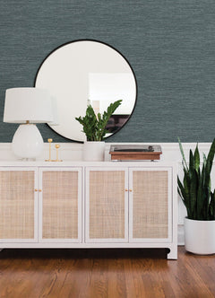 A modern sideboard with a white lamp, potted plants, and a record player sits beneath a round mirror on a wall adorned with Malin Faux Grasscloth Wallpaper by York Wallcoverings. The floor is wooden.