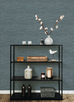 A black metal shelf displays décor items—a vase with cotton stems, candles, a wooden box, a decorative bird, bottles, and patterned boxes—set against a York Wallcoverings Malin Faux Grasscloth Wallpaper in blue-gray and a wood floor.