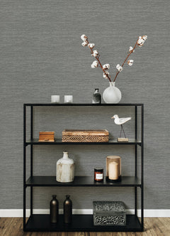 A black metal shelf displays decorative items—a vase with cotton stems, candles, boxes, bottles, a bird figurine, and a patterned box—set against a wall adorned with York Wallcoverings’ Malin Faux Grasscloth Wallpaper and a wooden floor.