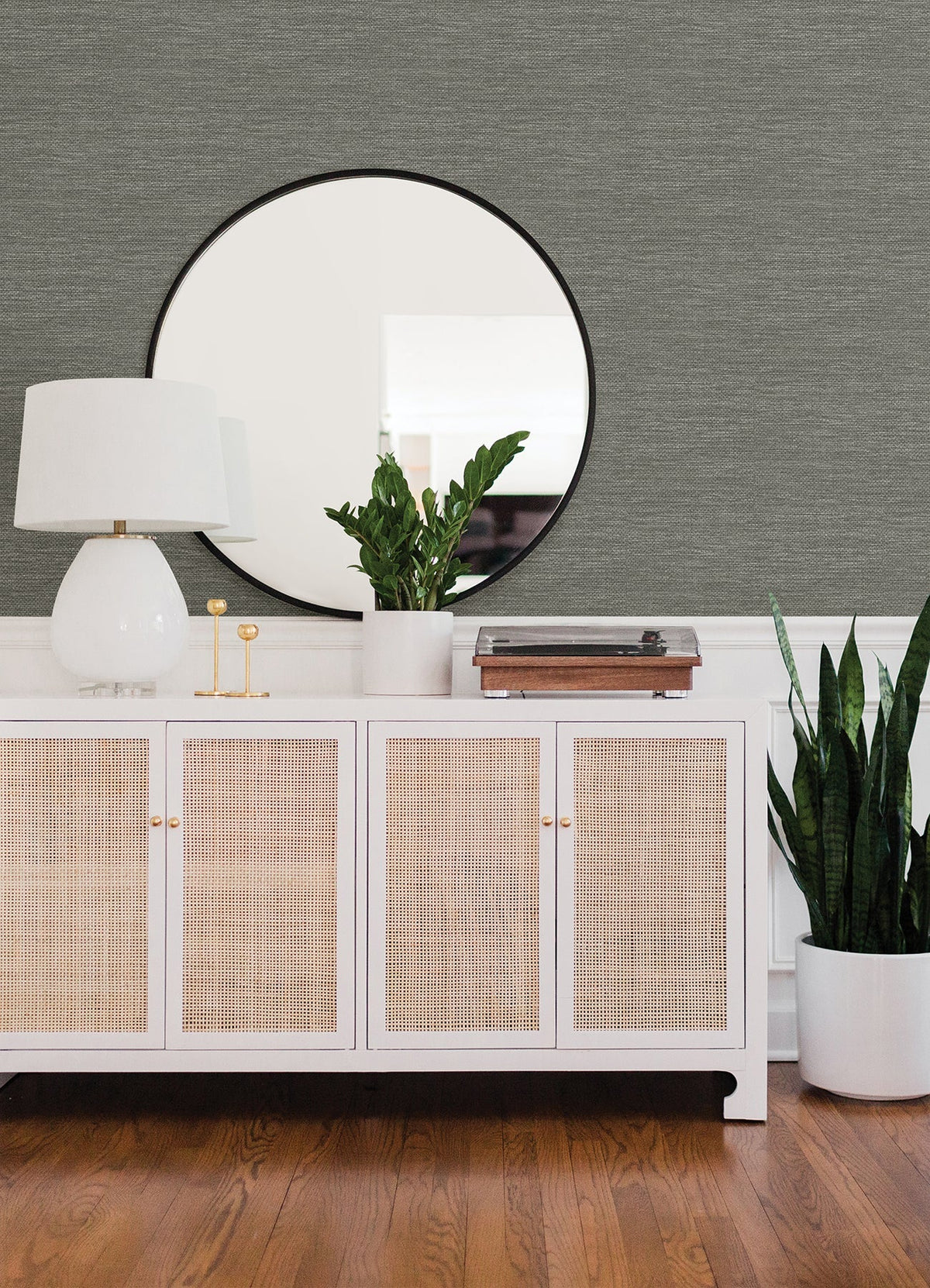 Adorned with Malin Faux Grasscloth Wallpaper by York Wallcoverings, the space features a white sideboard with wicker doors holding a lamp, potted plant, and record player beneath a round mirror. Beside the sideboard, a tall plant in a white pot rests on the hardwood floor.