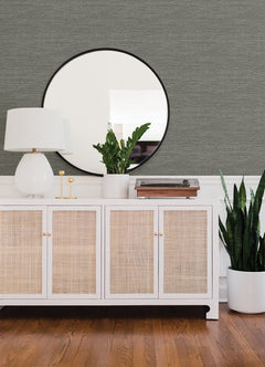Adorned with Malin Faux Grasscloth Wallpaper by York Wallcoverings, the space features a white sideboard with wicker doors holding a lamp, potted plant, and record player beneath a round mirror. Beside the sideboard, a tall plant in a white pot rests on the hardwood floor.