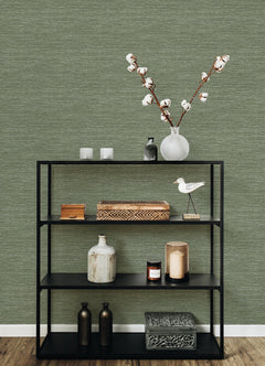 A black metal shelf holds decorative items—a vase with cotton stems, candles, boxes, bottles, a ceramic bird, and a patterned box—displayed against a wall covered in Malin Faux Grasscloth Wallpaper by York Wallcoverings.