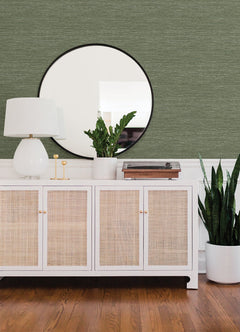 A white sideboard with cane doors sits on a wooden floor, topped with a white lamp, green plants, and decor. A round mirror hangs above against a wall covered in Malin Faux Grasscloth Wallpaper by York Wallcoverings.