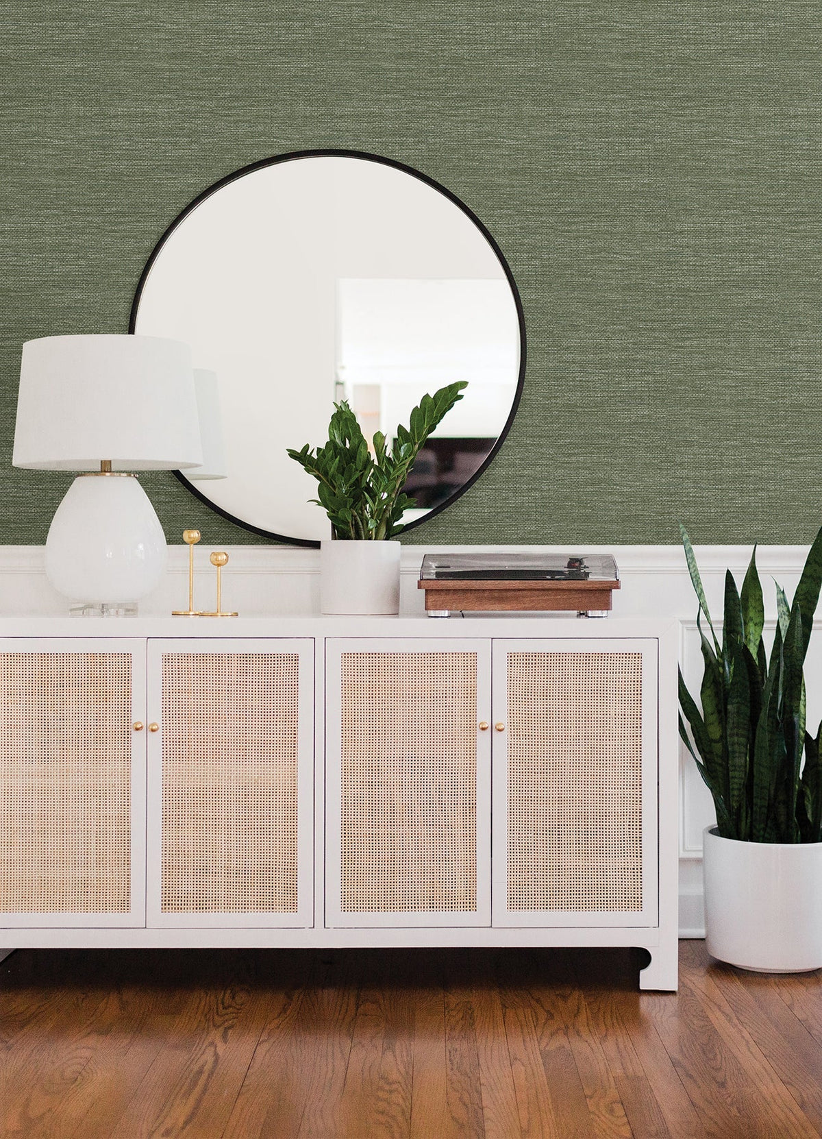 A white cabinet with cane doors holds a white lamp, small green plant, and record player. A large round mirror hangs above, reflecting the room’s Malin Faux Grasscloth Wallpaper by York Wallcoverings. A tall potted plant sits on the wooden floor nearby.