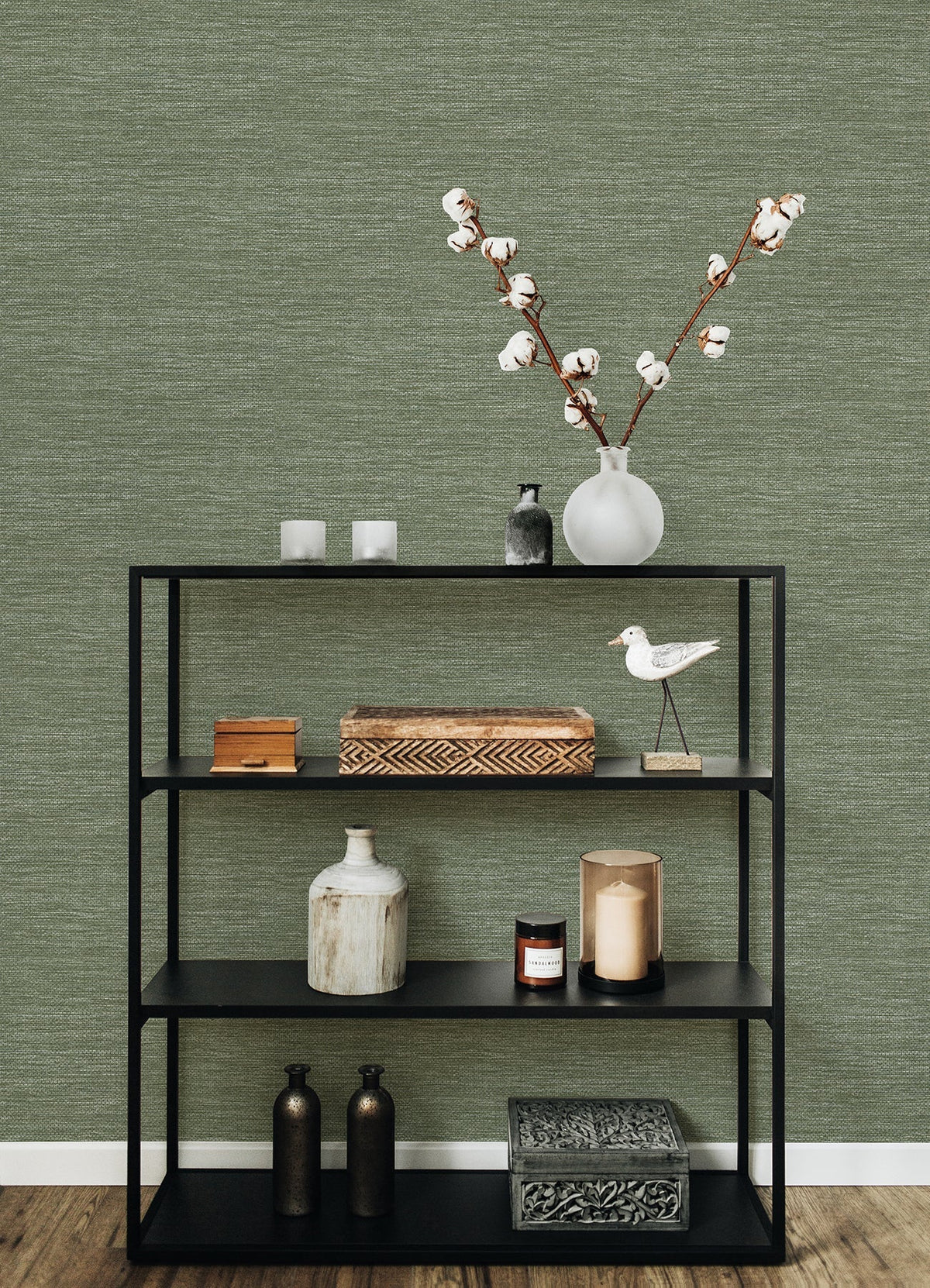 A black shelving unit holds decorative objects—a vase with cotton stems, candles, boxes, bottles, and a white bird figurine—set against a wooden floor and a textured green wall covered in Malin Faux Grasscloth Wallpaper by York Wallcoverings.