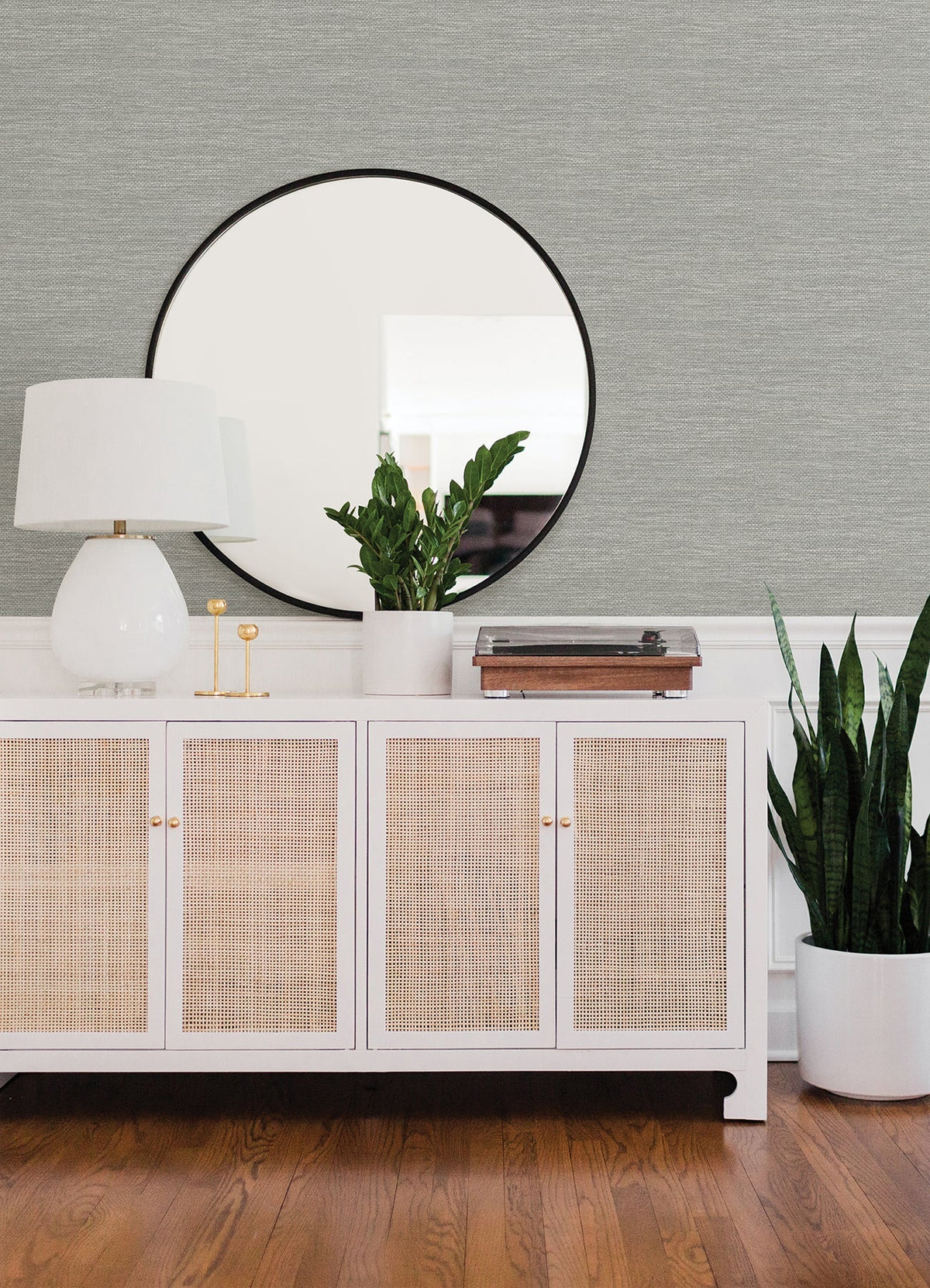A white sideboard with cane doors holds a white lamp, green potted plant, and record player. A round mirror hangs above, reflecting part of the room’s Malin Faux Grasscloth Wallpaper by York Wallcoverings. A tall plant sits beside the sideboard on a hardwood floor.